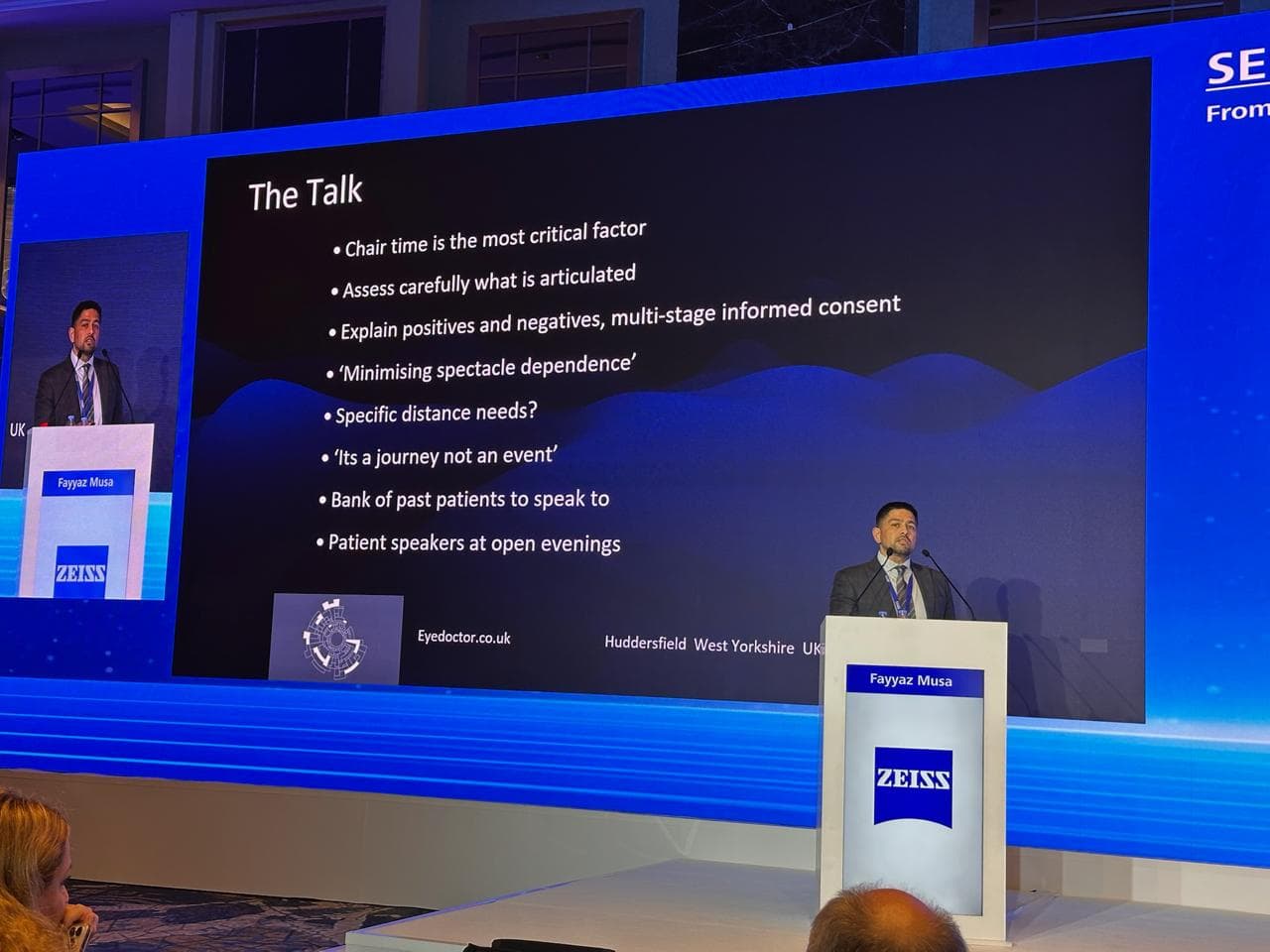 Mr Fayyaz Musa speaking at a ZEISS lectern, his presentation slide headed The Talk on the screen behind him
